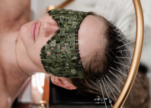 A young woman is lying on a spa table, enjoying a head spa treatment. She wears a soft green eye mask, and her posture exudes relaxation.