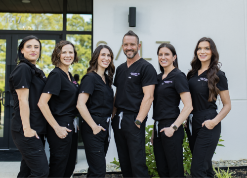 A group photo of the providers at Salt Spa and Wellness Center standing outside the medspa. They are smiling warmly and dressed in matching black scrubs, exuding professionalism and friendliness.